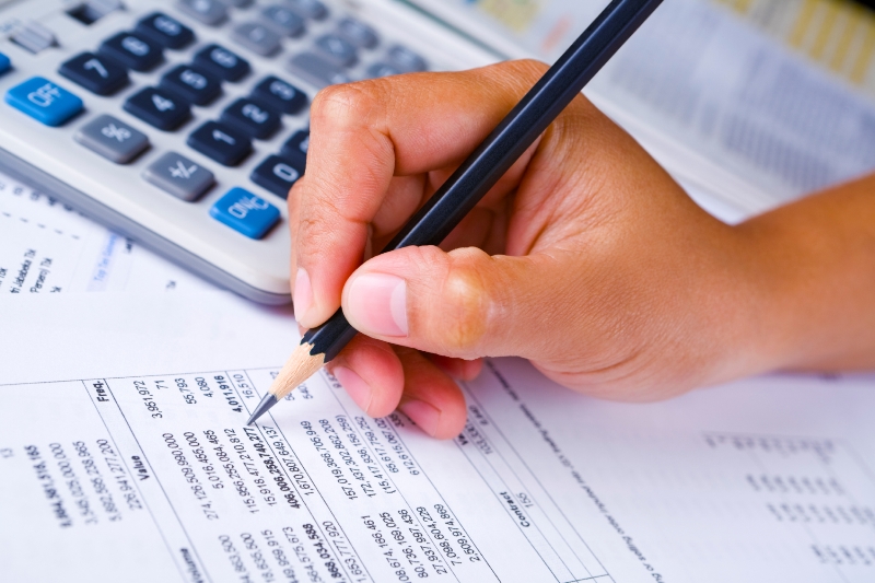 a Hand holding a pencil checking financial report, the calculator and newpaper are in the background.