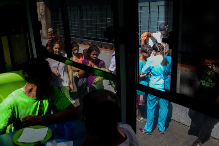 A volunteer doctor talks to a homeless person outside the PanaBus (FriendlyBus) while two homeless men eat a meal on the bus in Caracas on November 27, 2017. Monday through Friday, the PanaBus (FriendlyBus) makes its way around the Venezuelan capital offering homeless people a shower, clean clothes, a meal, a health check-up and advice to help them out of their plight - a beacon of hope and solidarity in the midst of economic and political crisis and severe shortages of food and medicines. / AFP PHOTO / FEDERICO PARRA / TO GO WITH AFP STORY by MARGIONI BERMUDEZ